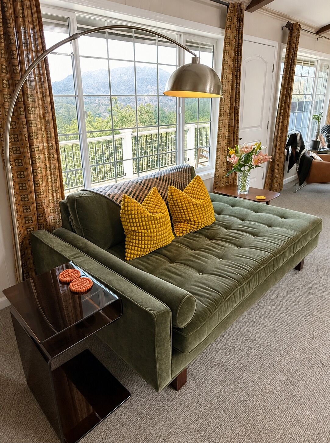 Un sofá verde con almohadones amarillos, una mesita de noche de madera oscura y un ventanal con vistas a zonas verdes y montañas.