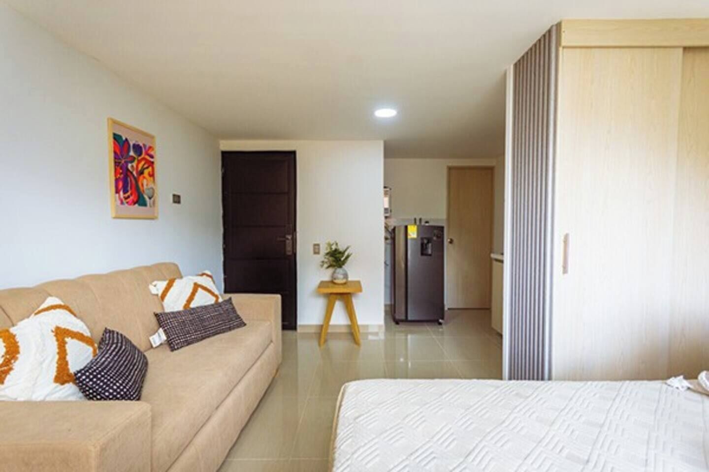A hotel room with a bed, a sofa, a small table with a plant, a refrigerator, and a door leading to another room.