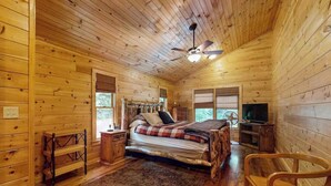 Ein Schlafzimmer einer Holzhütte mit Bett, Deckenventilator, Fernseher und Fenster.