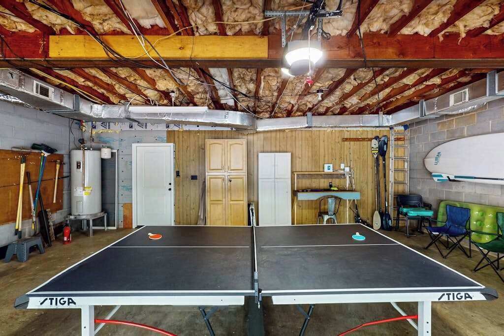 A ping pong table in a basement with insulation on the ceiling, a water heater, and various sports equipment.