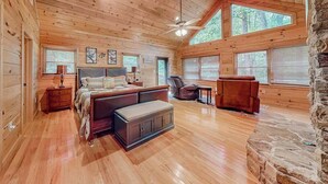 Ein Schlafzimmer in einer Blockhütte mit einem Bett, einem Sessel, einem kleinen Tisch und einem Kamin aus Stein.