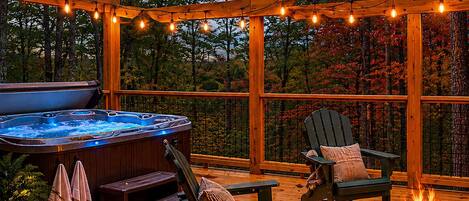 A wooden deck with a hot tub, two Adirondack chairs, a fire pit, and string lights.