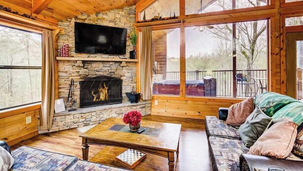Een gezellig interieur van een chalet met een stenen openhaard, houten wanden, een televisie erboven en een groot raam met uitzicht op de natuur.