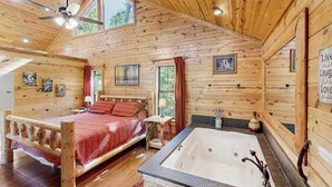 Una habitación tipo cabaña de madera con una cama de madera, un ventanal grande, un ventilador de techo y una bañera.
