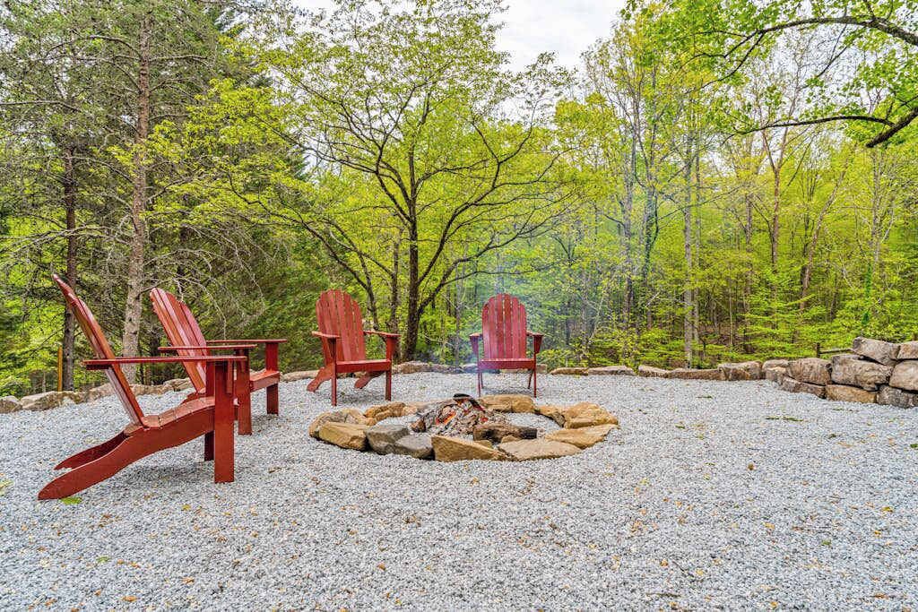 A forested area with a fire pit and three Adirondack chairs.