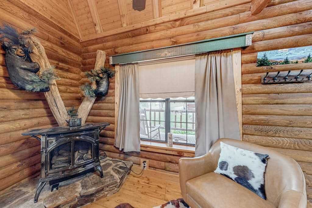 A log cabin interior with a wooden stove, a window with curtains, and a leather sofa.