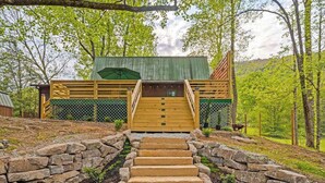 A wooden staircase leading to a cabin with a green roof, surrounded by trees and a stone wall.