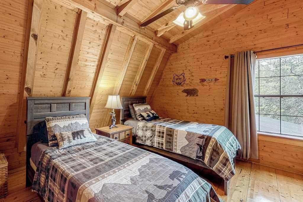 A wooden cabin bedroom with two beds, a ceiling fan, and a window with curtains.