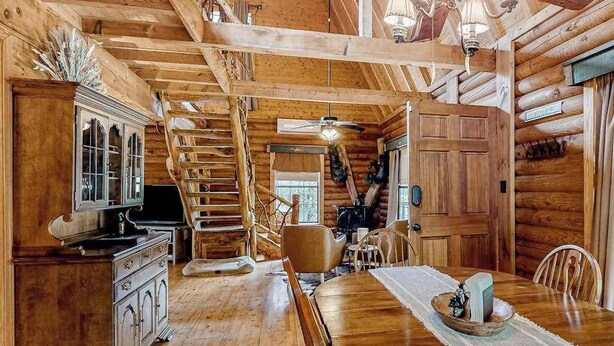 A log cabin interior with a dining table, chairs, a staircase, and a kitchen area.