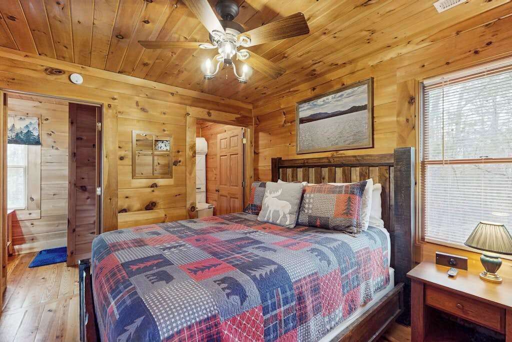 Un dormitorio de cabaña de madera con una cama, un ventilador de techo, una ventana con persianas y un cuadro en la pared.