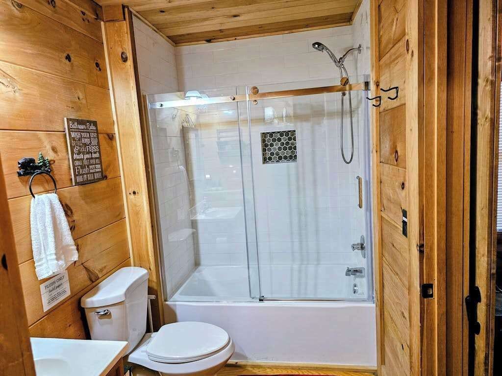 Un baño de cabaña de madera con ducha acristalada, inodoro y lavamanos.