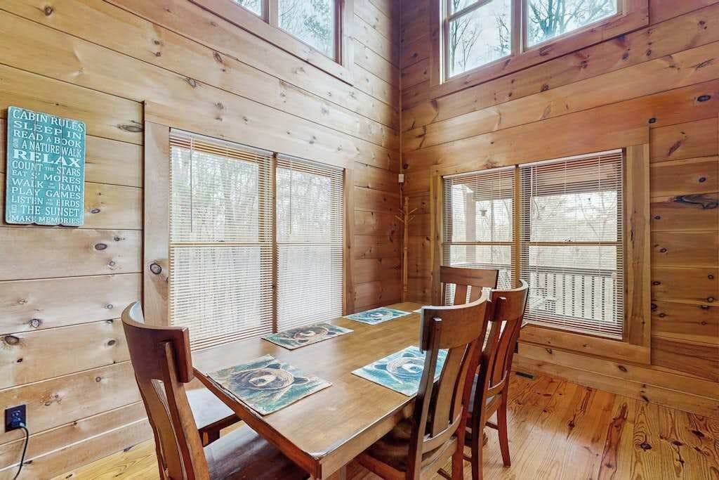 A wooden cabin interior with a dining table, chairs, and large windows with blinds.