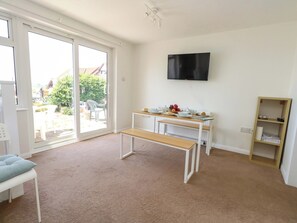 A room with a sliding glass door, a TV mounted on the wall, a wooden table with a bench, and a bookshelf. - Bella Rosa (Weymouth)