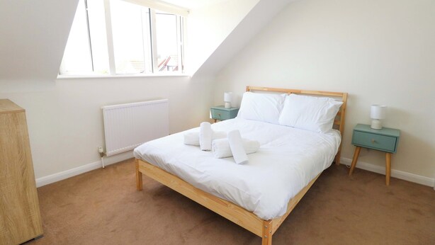 Quarto com teto inclinado, cama de madeira, radiador, janela e mesa de cabeceira com abajur.