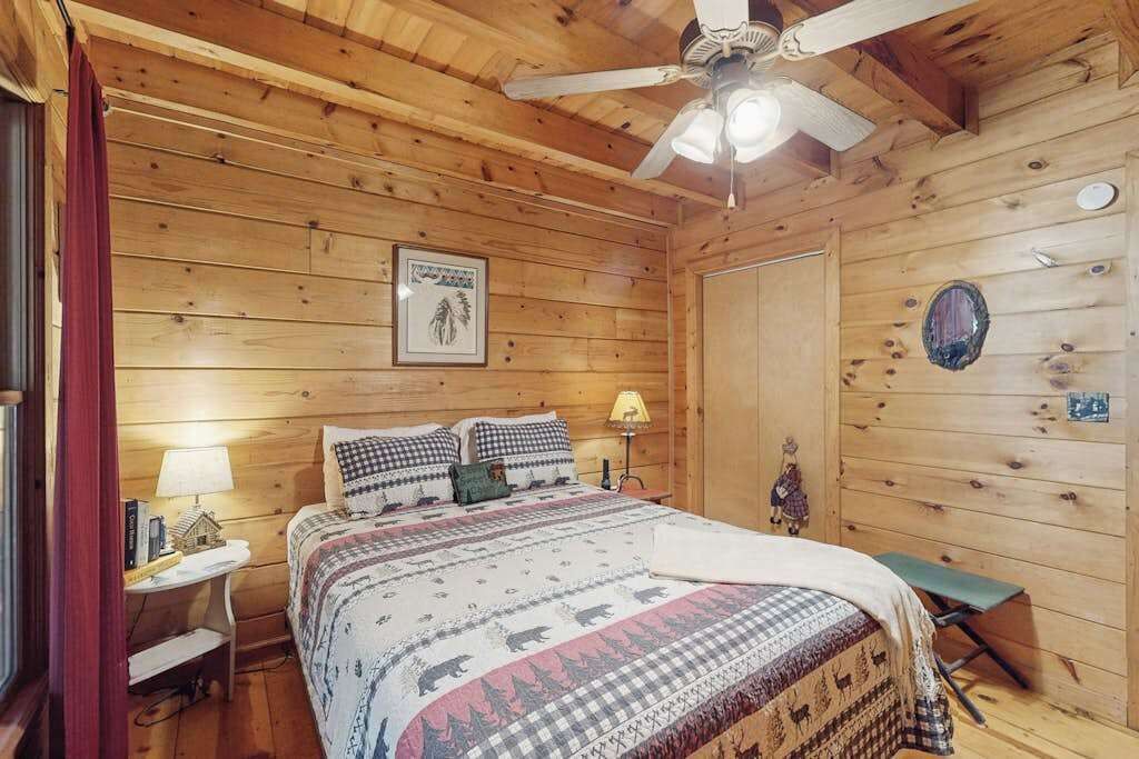 A wooden cabin bedroom with a bed, a nightstand, a desk, a chair, a ceiling fan, and a door.