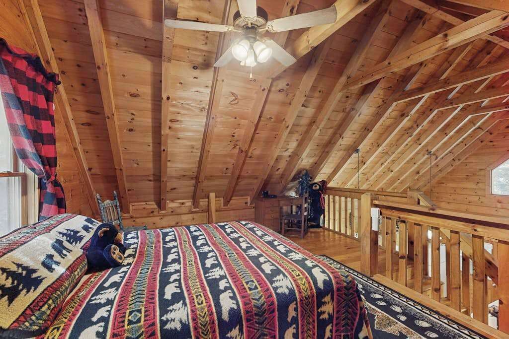 Une cabane en bois avec un plafond en pente, un lit recouvert d’une couverture à motifs, un ventilateur de plafond et une rambarde en bois.