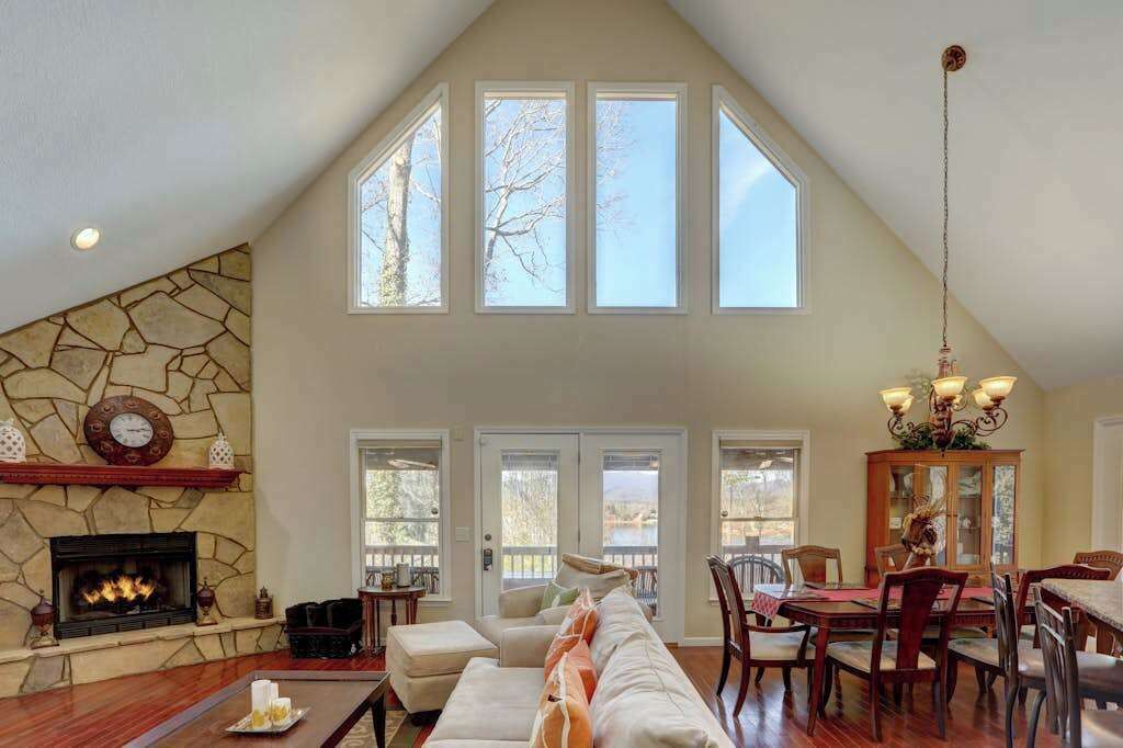 A spacious living room with a stone fireplace, a high ceiling with skylights, a dining area with wooden furniture, and a view of the outdoors.