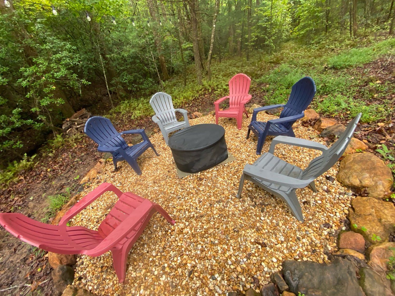 Disposição circular de assentos com cadeiras Adirondack em várias cores, uma mesa redonda preta e piso com pedriscos.