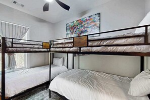 Een slaapkamer met een stapelbed, een raam, een plafondventilator en een kleurrijk schilderij aan de muur.