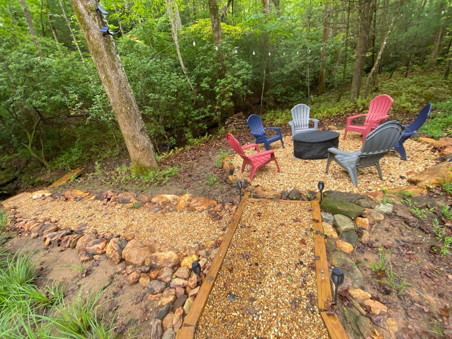 An outdoor seating area with Adirondack chairs and a fire pit surrounded by a gravel path and natural forest backdrop.