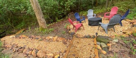An outdoor seating area with Adirondack chairs and a fire pit surrounded by a gravel path and natural forest backdrop.