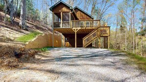 Une cabane en bois à deux étages, dotée d’un balcon, d’une allée en gravier et située sur le versant d’une colline boisée.