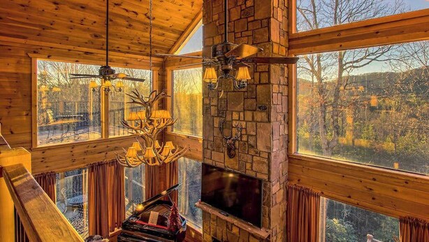 Interior de una cabaña con chimenea de piedra, ventanas amplias y un ventilador de techo.