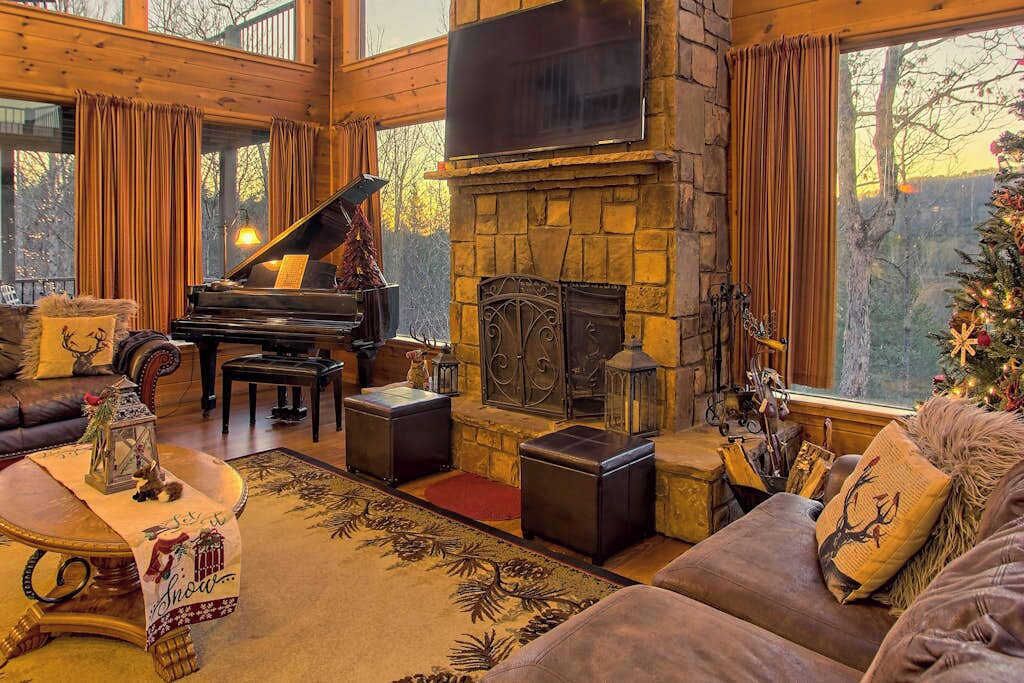 A cozy living room with a grand piano, a stone fireplace, and a large window overlooking a forest.