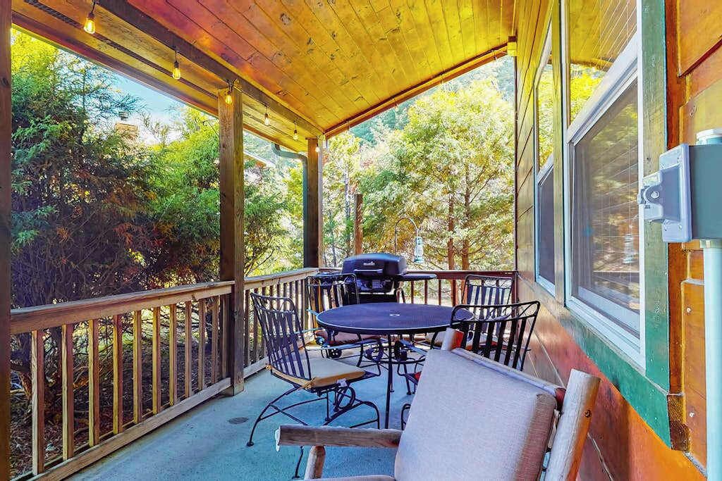 A covered outdoor patio with a table and chairs, a grill, and a view of trees.