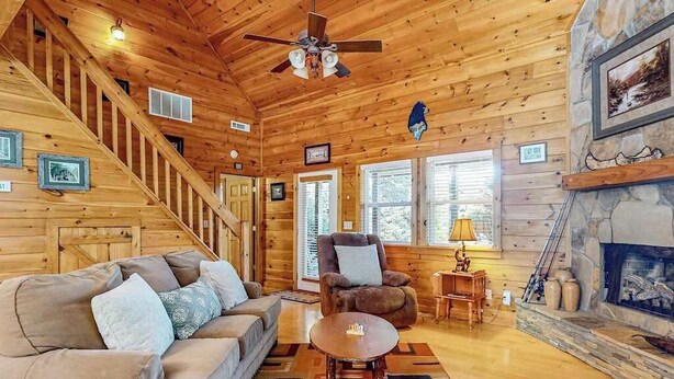 L’intérieur d’une cabane en rondins, avec un escalier en bois, un canapé, un fauteuil, une cheminée et un ventilateur de plafond.