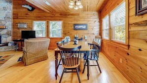 L’intérieur d’un chalet en bois comprend une table à manger, des chaises, un téléviseur et des tableaux encadrés accrochés aux murs.