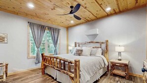 Eine Holzbalkendecke, ein Bett mit Kopfteil, ein Nachttisch, ein Fenster mit Vorhängen und ein Deckenventilator.
