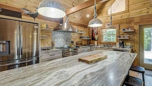 A wooden cabin interior with a large kitchen island, stainless steel appliances, and hanging pendant lights. - The Mountain Chapter (Blairsville)