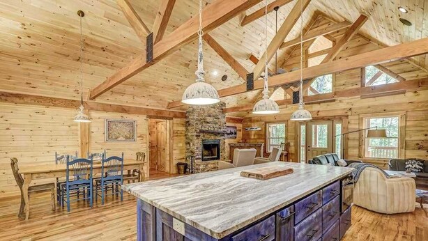 A spacious log cabin interior with a large wooden dining table, blue chairs, a stone fireplace, and a kitchen island with drawers. - The Mountain Chapter (Blairsville)
