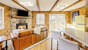 Un interno accogliente di una baita con camino in pietra, pareti in legno, un divano in pelle, zona pranzo e cucina con frigorifero.