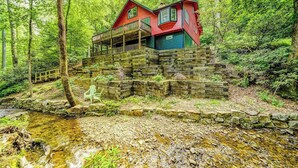 Eine Holzhütte mit einer Holzterrasse und einer Steintreppe, die zu einem roten und blauen Haus im Wald führt.