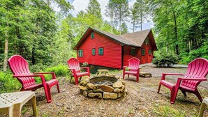 Eine rote Holzhütte mit einem Steinkamin und vier roten Adirondack-Stühlen in einer Waldlandschaft.