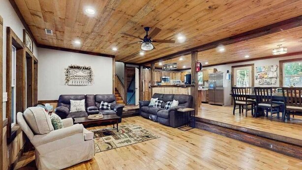 Amplia sala de estar con techo de madera, un conjunto de sofás, un comedor con mesa y sillas, y una cocina al fondo.