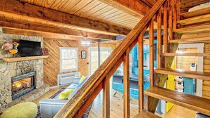 Interno di una baita in legno con camino in pietra, una scala e un soggiorno con divano e cuscini.