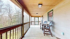 A covered porch with a wooden railing, a table with chairs, and a view of trees through the windows. - Our Mountain Blessing (Blairsville)