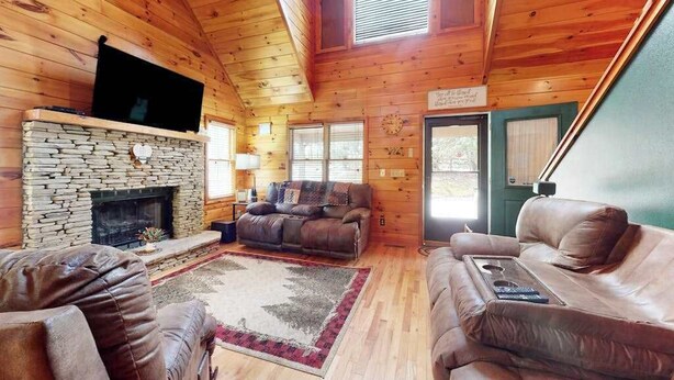 A log cabin interior with a stone fireplace, leather sofas, a flat-screen TV, and a large window. - Our Mountain Blessing (Blairsville)