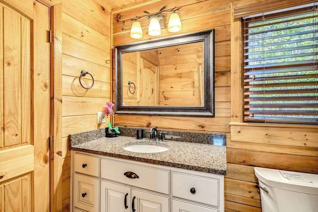 A wooden cabin bathroom with a granite countertop sink, a large mirror, and a window with blinds.