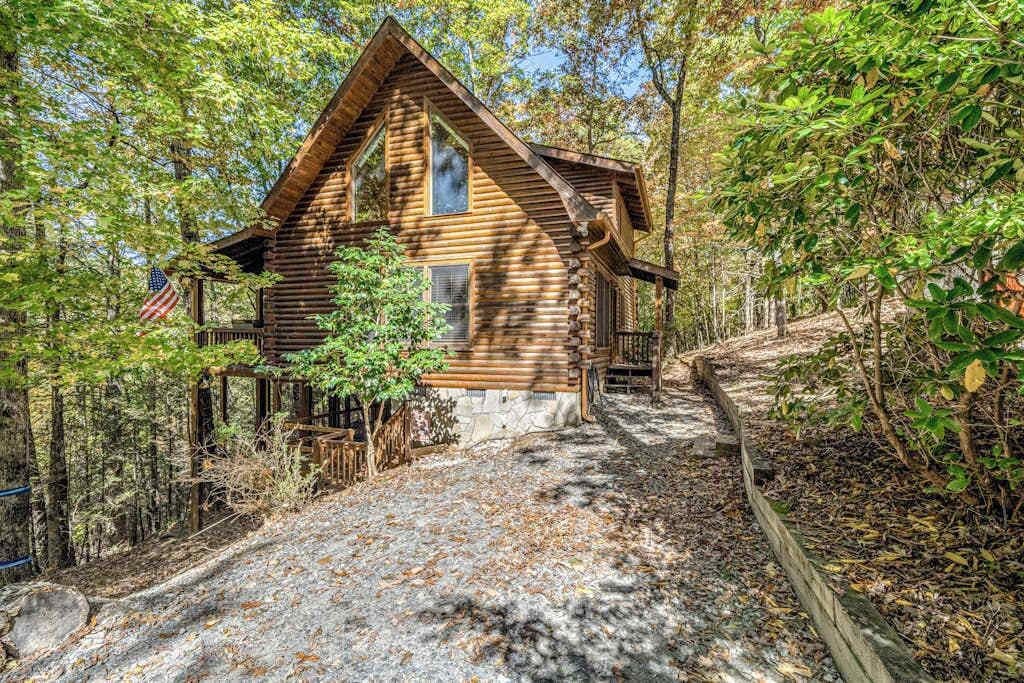 A log cabin with a stone foundation, surrounded by trees and a gravel path leading to the entrance.
