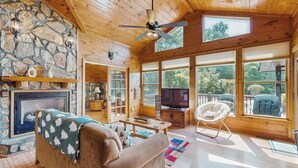 A cozy cabin interior with a stone fireplace, wooden ceiling, large windows, a television, and a comfortable seating area. - Time Out Blue Ridge - Hot tub (Blue Ridge)