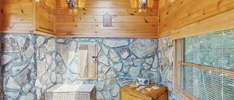 A cabin interior with wooden walls, a stone wall, a window with blinds, a basket, a wooden box, and two wall-mounted lanterns.