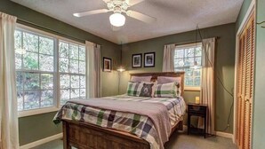 Une chambre à coucher avec un lit en bois, deux fenêtres avec des rideaux, un ventilateur de plafond et une table de chevet avec une lampe.