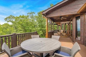 A wooden deck with a round table and wicker chairs, surrounded by a wooden railing, overlooking a forested area. - Endless Views Retreat (Mineral Bluff)