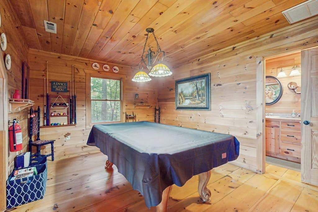 A wooden cabin interior with a pool table, wooden walls, a door with a mirror, and a shelf with items.