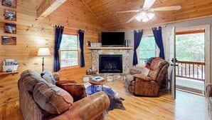 Interior de una cabaña de madera con chimenea, muebles de cuero, televisor y ventilador de techo.