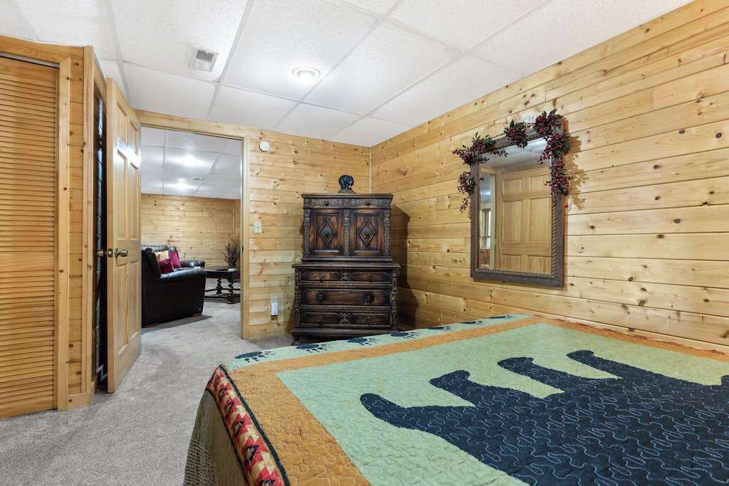 Une pièce aux murs en bois, avec un lit recouvert d’une couvre-lit à motifs, une commode, un miroir et une porte donnant sur une autre pièce.